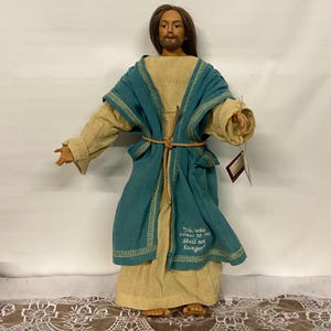 May include: A handcrafted doll depicting a figure in a long, tan robe with a teal overcoat. The overcoat is trimmed with gold stitching and tied with a brown cord. The text "He who comes to Me Shall not hunger" is embroidered on the coat.