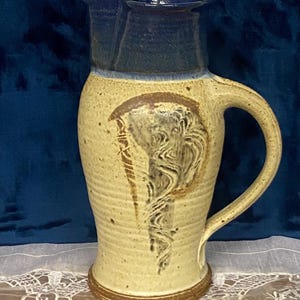 May include: A handmade ceramic pitcher with a tan body and a dark blue rim. The pitcher features a large handle and a decorative swirl design in brown and gray. The base is a darker brown. The background is a dark blue.