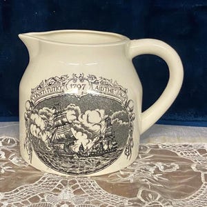 May include: A cream-colored ceramic pitcher featuring a black and white transfer print of two sailing ships on the ocean. The design includes an ornate border with the text CONSTITUTION 1797 AID THE JAVA arched above the nautical illustration.