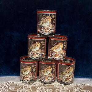 May include: Six clear drinking glasses with a red band around the top. Each glass features a detailed illustration of a bird with the words "Free winged soul" in gold lettering. The glasses are stacked in a pyramid shape.