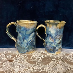 May include: Two ceramic mugs with a tall, cylindrical shape. The mugs feature a blue and tan glaze with a dripped effect. Each mug has a large, curved handle. The mugs are displayed on a white lace tablecloth.