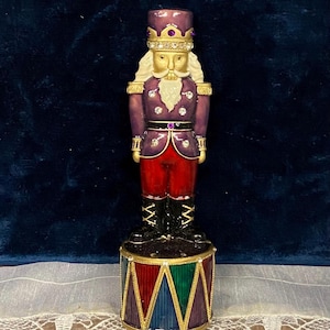 May include: A decorative Christmas stocking holder shaped like a nutcracker soldier. The figure is dressed in a purple jacket, red pants, and a gold-trimmed hat. The base is a drum with red, green, and blue panels.