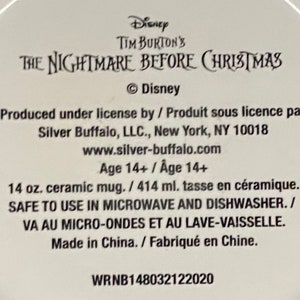 Op de afbeelding: Een witte keramische mokbodem met zwarte tekst die Disney Tim Burton's The Nightmare Before Christmas leest. Het bevat productdetails zoals een inhoud van 414 ml, informatie over magnetron- en vaatwasserbestendigheid en de fabrikant Silver Buffalo, LLC.
