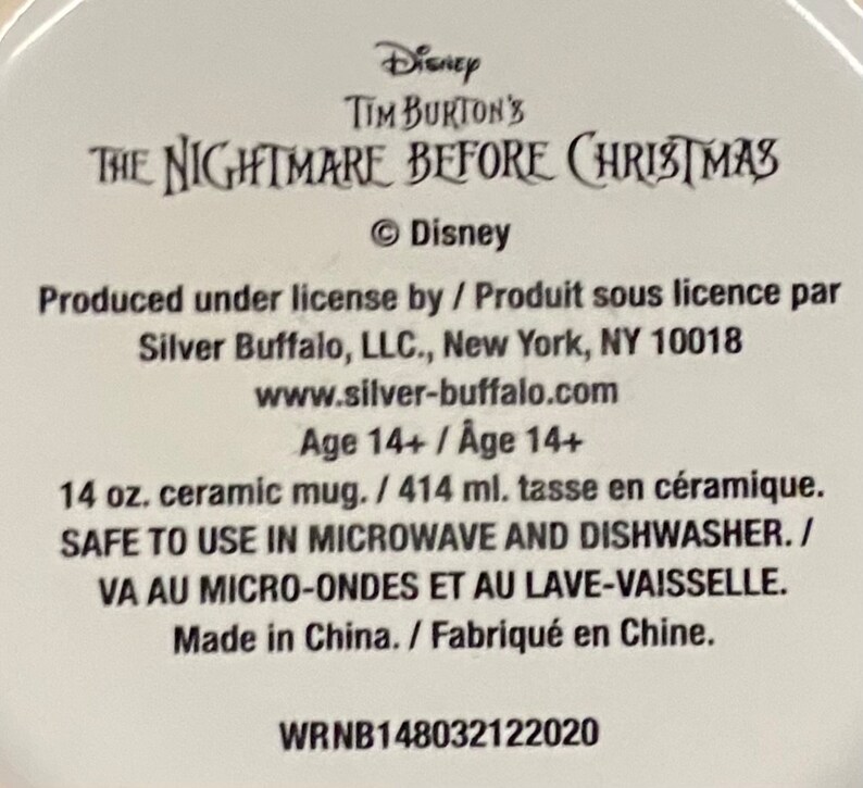 Op de afbeelding: Een witte keramische mokbodem met zwarte tekst die Disney Tim Burton's The Nightmare Before Christmas leest. Het bevat productdetails zoals een inhoud van 414 ml, informatie over magnetron- en vaatwasserbestendigheid en de fabrikant Silver Buffalo, LLC.