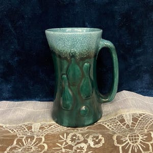 May include: A vintage ceramic mug with a teal rim and handle, transitioning to a dark green body. The mug features a textured surface on the rim and decorative teardrop-shaped accents. The mug is displayed on a white lace cloth.