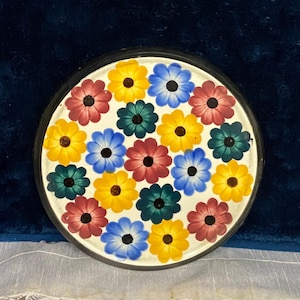 May include: A round, decorative plate with a black rim and a white center. The center is adorned with a pattern of colorful flowers in shades of red, yellow, blue, and green. The flowers are evenly spaced across the surface.