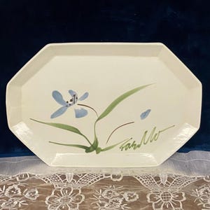 May include: Cream-colored, octagonal ceramic serving platter with a hand-painted blue flower and green leaves. The platter has a signature in brown ink. A decorative piece for serving food.
