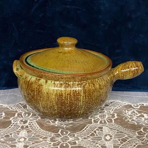 May include: A ceramic soup tureen with a lid and handle. The tureen is a mottled brown and tan color with a green rim on the lid. The tureen is sitting on a white lace tablecloth.