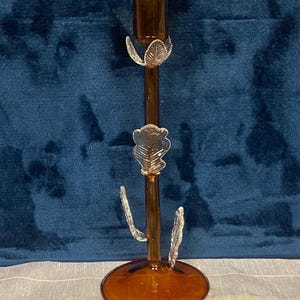 May include: A decorative amber glass candlestick with leaf accents. The candlestick has a round base and a slender stem with three leaf-shaped embellishments. The top is designed to hold a single candle. The background is a dark blue velvet.