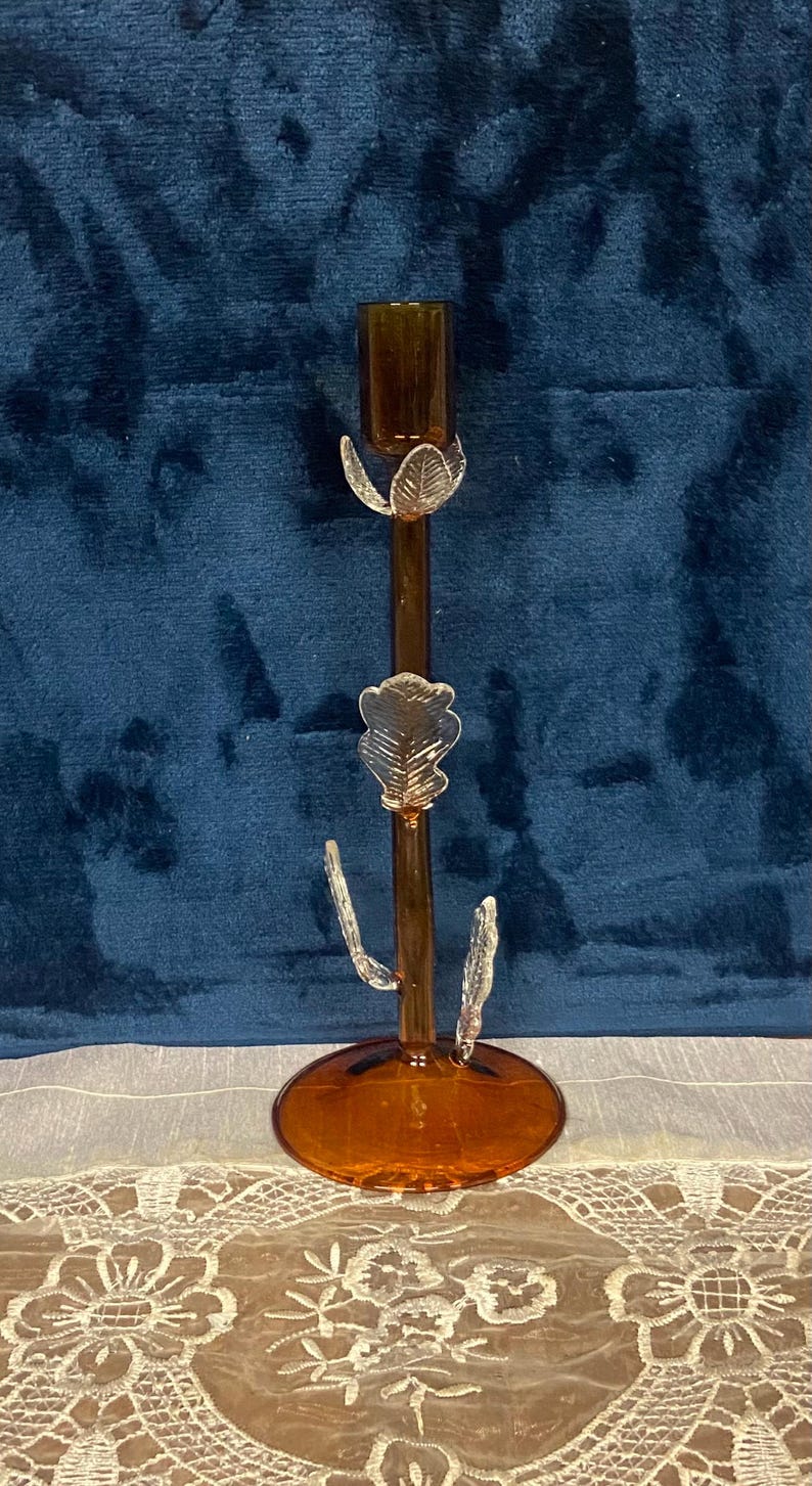 May include: A decorative amber glass candlestick with leaf accents. The candlestick has a round base and a slender stem with three leaf-shaped embellishments. The top is designed to hold a single candle. The background is a dark blue velvet.