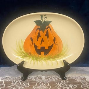 May include: An oval-shaped cream-colored ceramic platter featuring a hand-painted orange jack-o'-lantern with a black carved face and green leaves at the top. The pumpkin sits atop a base of painted green grass strokes on the platter surface.