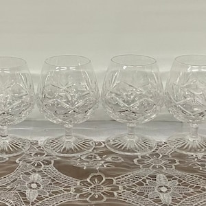 May include: Four clear crystal brandy snifters with intricate cut glass patterns. The glasses have a rounded bowl shape, a short stem, and a wide, decorative base. They are arranged in a row on a lace tablecloth.