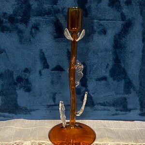 May include: A decorative amber-colored glass candlestick holder with clear glass accents. The candlestick has a round base, a slender stem, and clear glass leaf-like details. The top is a small cup to hold a candle.