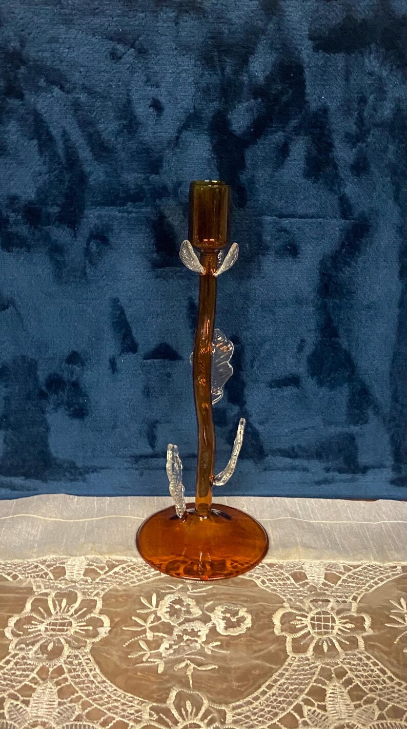 May include: A decorative amber-colored glass candlestick holder with clear glass accents. The candlestick has a round base, a slender stem, and clear glass leaf-like details. The top is a small cup to hold a candle.