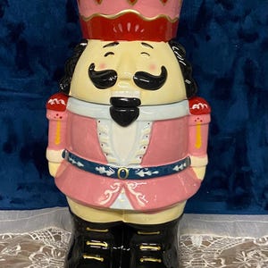 May include: A ceramic nutcracker cookie jar featuring a pink uniform with a red crown, black boots, and a gold finial. The figure has a cream-colored face, black mustache, and beard, with a blue belt featuring white floral accents and gold details.