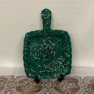 May include: A dark green ceramic serving board with a handle. The board features a raised floral design with leaves and flowers. The board is rectangular with a scalloped edge.