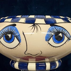 May include: A decorative ceramic bowl with a whimsical face design. The bowl features a cream-colored base with a painted face, including blue eyes, eyelashes, and a pink mouth. The rim is checkered blue and white, and the base has blue and white stripes.