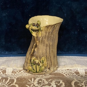 May include: A ceramic vase with a textured, tree-trunk-like exterior. Two green frog figurines are attached to the vase, one at the top and one at the base. The interior of the vase is a light cream color.