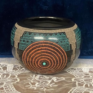 May include: A round, black ceramic bowl with intricate designs. The bowl features a central orange spiral pattern with a turquoise center, surrounded by geometric teal and gray designs. The bowl sits on a white lace surface.