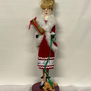 May include: A decorative Christmas figurine of a woman in a red coat with white fur trim, holding a wrapped gift and a green leash attached to a small dog figurine. The figurine stands on a red base.