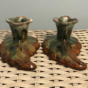 May include: A pair of ceramic candle holders featuring a glossy glaze in shades of deep brown and forest green. Each holder has a wide, leaf-shaped base and a flared, flower-shaped top opening designed to securely hold a standard taper candle.