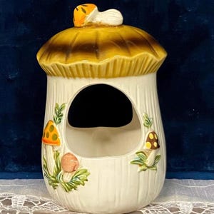 May include: A ceramic mushroom-shaped container with a large opening. The container is primarily white with a brown and yellow mushroom cap lid. Decorative mushrooms and foliage are painted on the sides. The container is sitting on a white lace tablecloth.