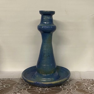 May include: A blue ceramic candlestick with a wide base and a tall, slender stem. The candlestick has a glazed finish with a subtle green hue.