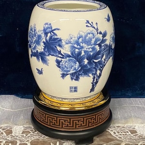 May include: A white ceramic vase with blue floral peony designs and small butterfly accents. The vase sits on a black wooden stand with a gold trim and a decorative brown base. The vase is a decorative home decor item.
