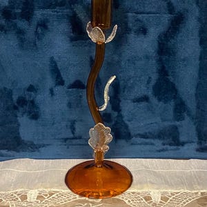 May include: Amber-colored glass candlestick holder with a curved stem and leaf-like accents. The base and candle cup are round. The candlestick is on a white lace tablecloth against a blue background.
