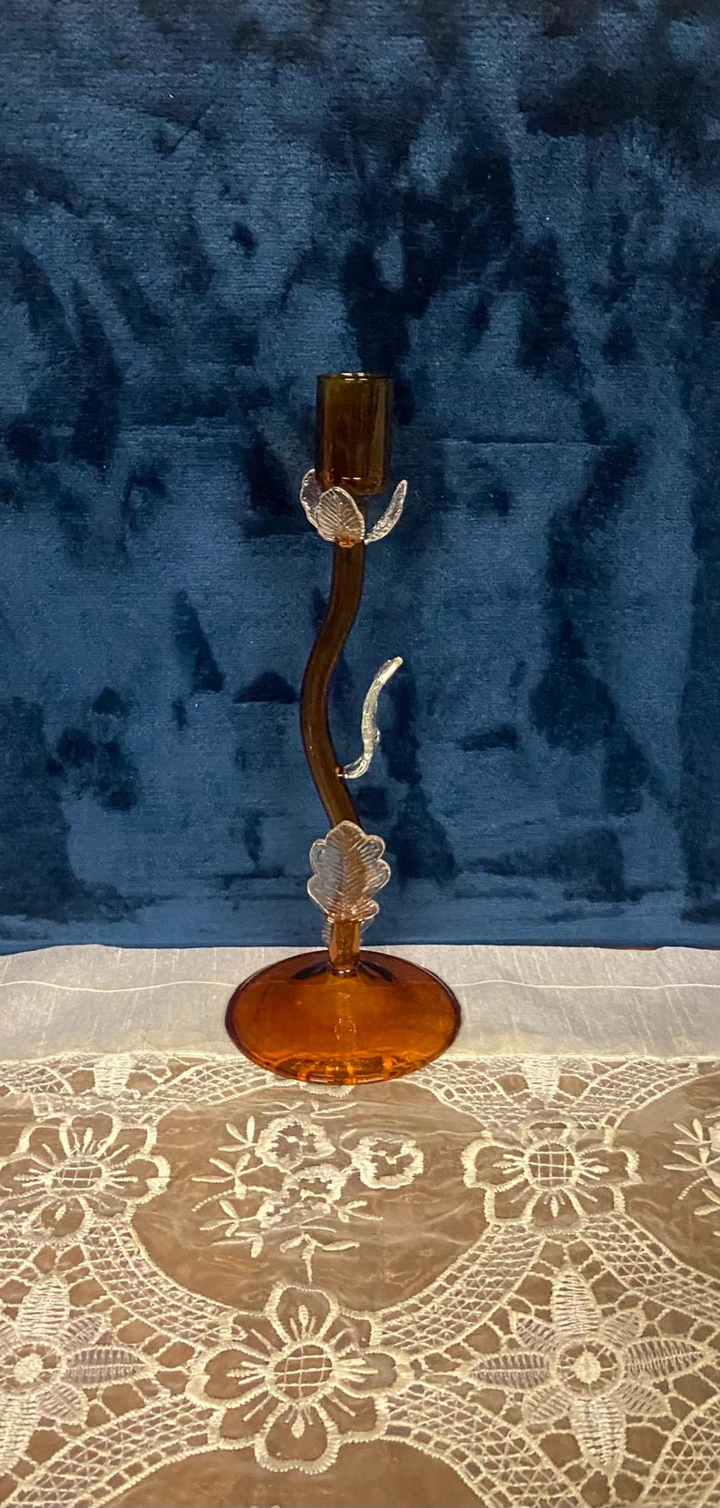 May include: Amber-colored glass candlestick holder with a curved stem and leaf-like accents. The base and candle cup are round. The candlestick is on a white lace tablecloth against a blue background.