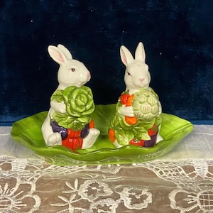 May include: A ceramic salt and pepper set featuring two white bunny figurines, each holding a bundle of vegetables. The bunnies sit on a green, leaf-shaped dish. The vegetables include red, purple, and green accents.
