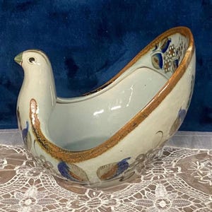 May include: A ceramic bird-shaped bowl with a cream-colored glaze, featuring hand-painted blue and brown floral motifs and a distinct brown rim. The vessel has a smooth, curved body with a raised head and tail, showcasing a decorative pattern.