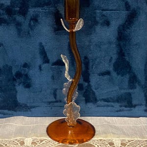 May include: A decorative amber-colored glass candlestick holder. The candlestick has a curved stem with clear leaf-shaped accents and a round base. The top is designed to hold a single candle. The background is a dark blue velvet.