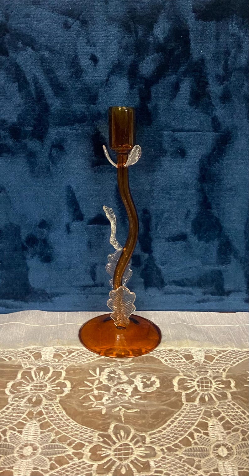 May include: A decorative amber-colored glass candlestick holder. The candlestick has a curved stem with clear leaf-shaped accents and a round base. The top is designed to hold a single candle. The background is a dark blue velvet.
