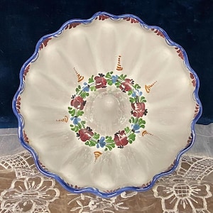 May include: A white ceramic serving bowl with a scalloped edge and a blue rim. The bowl features a floral design in the center with red, blue, and green accents. The bowl is approximately 12 inches in diameter.