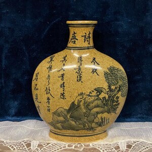 May include: A tan ceramic vase with black calligraphy and a painted scene of a man under a tree. The vase has a rounded body and a narrow neck. The artwork is in a traditional Asian style.