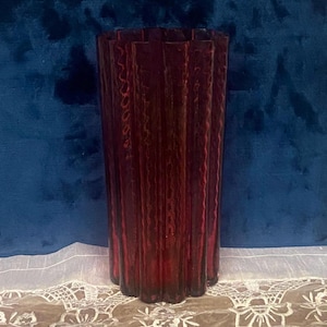 May include: A tall, fluted vase in a deep burgundy hue. The vase has a textured surface and a unique, vertical ribbed design. The vase is set against a dark blue backdrop and a white lace tablecloth.
