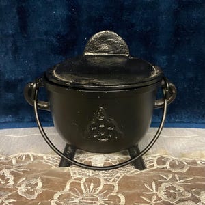 May include: A black cast iron cauldron with a matching lid, three short legs, and a curved metal handle. The front of the pot features a raised triquetra symbol. The surface has a matte finish, and the lid includes a small, semi-circular handle.