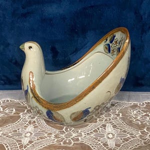 May include: A ceramic bird-shaped bowl with a white body, brown rim, and blue floral accents. The bird has a long, curved body with a head and beak at one end. The bowl is likely used for decorative purposes.