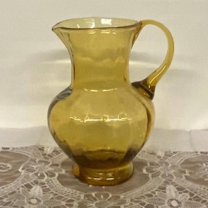 May include: Amber-colored glass pitcher with a rounded body, a flared rim, and a curved handle. The pitcher has a textured surface and a small base. The glass is transparent, allowing light to pass through. The pitcher is likely used for serving beverages.