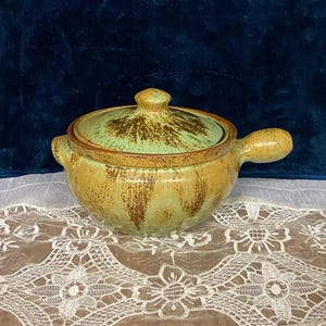 May include: A ceramic soup pot with a lid. The pot is a light yellow-brown color with green and brown accents. It has a handle on one side and a lid with a knob. The pot is sitting on a white lace tablecloth.