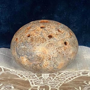 May include: A spherical ceramic flower frog with a mottled brown, tan, and grey stone-like glaze. The rounded object features multiple circular openings across its top surface, designed for arranging stems in a floral display or centerpiece.