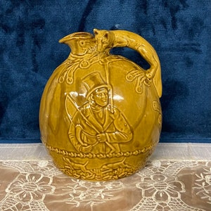 May include: A brown ceramic pitcher with a fox handle and a relief design of a man in a hat holding a stick. The pitcher is decorated with a rope design around the bottom.