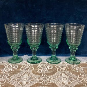 May include: Four green-tinted, ribbed-glass goblets with a classic design. The glasses have a rounded bowl, a stem, and a circular base. The goblets are arranged in a row on a white lace tablecloth, against a dark blue background.