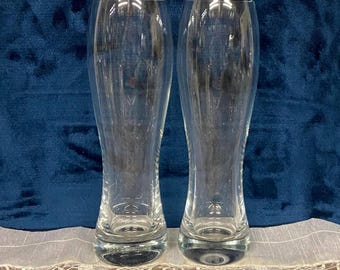 May include: Two clear, tall beer glasses with a slender, curved design. The glasses are empty and sit on a white lace tablecloth against a dark blue background. The glasses are made of clear glass.