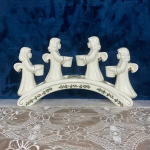 May include: A white ceramic Christmas candle holder featuring four angel figurines. Each angel holds a small cup. The arched base is decorated with green holly and red berries. Gold accents are present on the wings and cups.