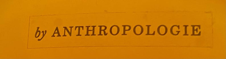 May include: The image displays the text "by ANTHROPOLOGIE" in a dark brown font, set against a solid orange background. The text is enclosed within a rectangular frame, suggesting a label or branding element.