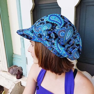 May include: A blue paisley print hat with a curved brim. The hat features a black background with blue, teal, and white paisley designs. The hat is made of fabric and is worn on the head.