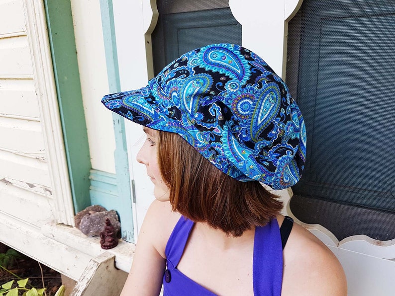 May include: A blue paisley print hat with a curved brim. The hat features a black background with blue, teal, and white paisley designs. The hat is made of fabric and is worn on the head.