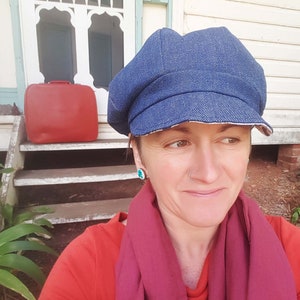 May include: A blue denim newsboy cap with a white brim. The cap is worn by a person wearing a red shirt and a burgundy scarf. A red suitcase sits on the steps behind the person.